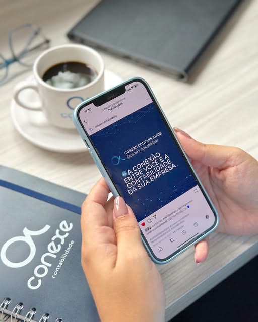 Mesa de trabalho com caderno da Conexe Contabilidade, café e celular aberto no perfil da Conexe
