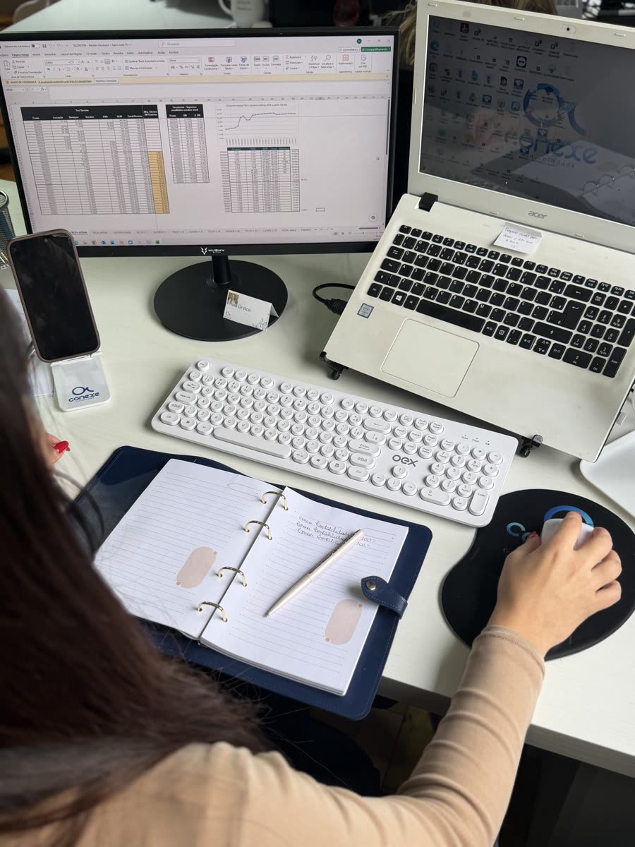 Profissional da Conexe trabalhando com planilhas no computador e agenda aberta sobre a mesa